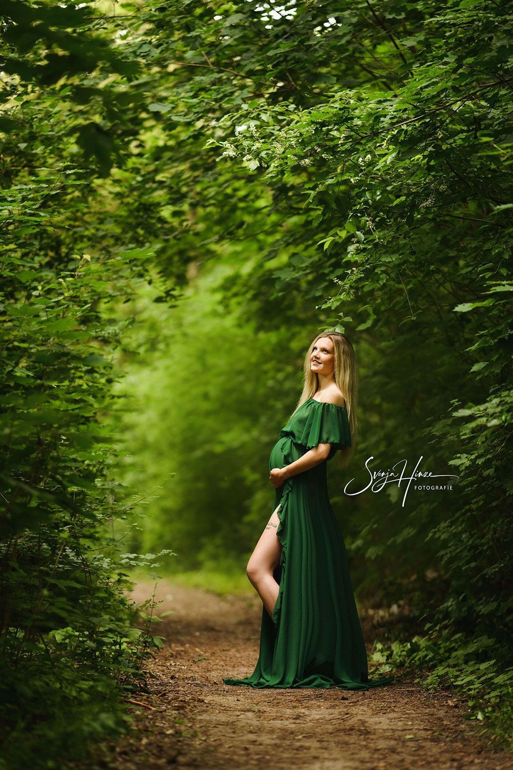 Eine schwangere Frau in einem grünen Kleid steht auf einem Waldweg.