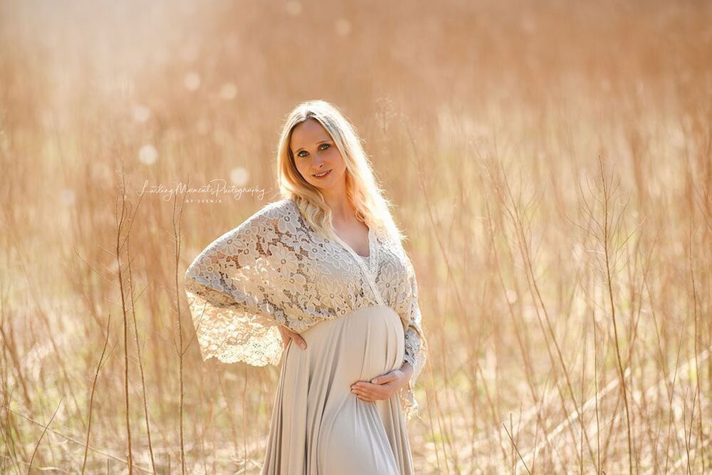 Eine schwangere Frau in einem weißen Spitzenkleid steht auf einem Feld und hält ihren Bauch.