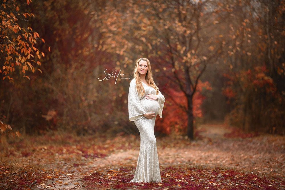 Eine schwangere Frau in einem weißen Kleid steht in einem Wald und hält ihren Bauch.