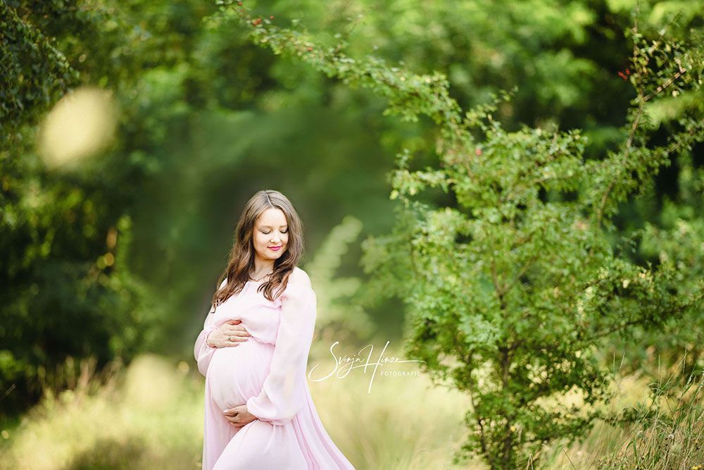 Eine schwangere Frau in einem rosa Kleid steht auf einem Feld und hält ihren Bauch.