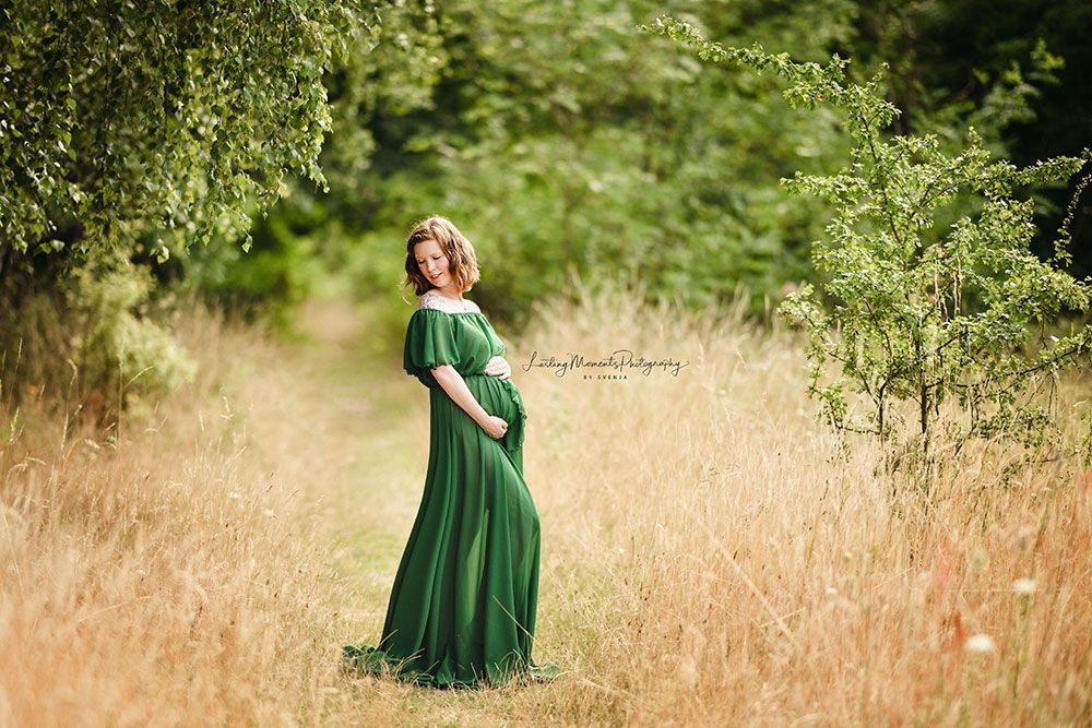 Eine schwangere Frau in einem grünen Kleid steht auf einem Feld und hält ihren Bauch.
