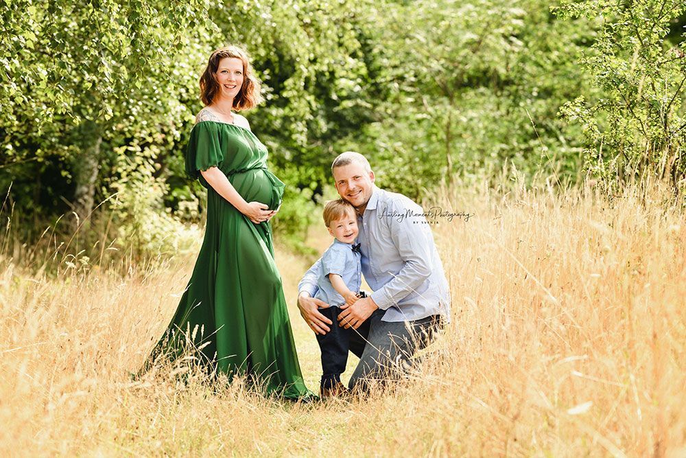 Eine schwangere Frau in einem grünen Kleid posiert mit ihrem Mann und ihrem Sohn auf einem Feld für ein Foto.