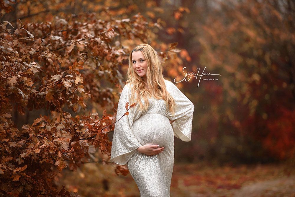 Eine schwangere Frau in einem weißen Kleid steht vor einem Baum und hält ihren Bauch.