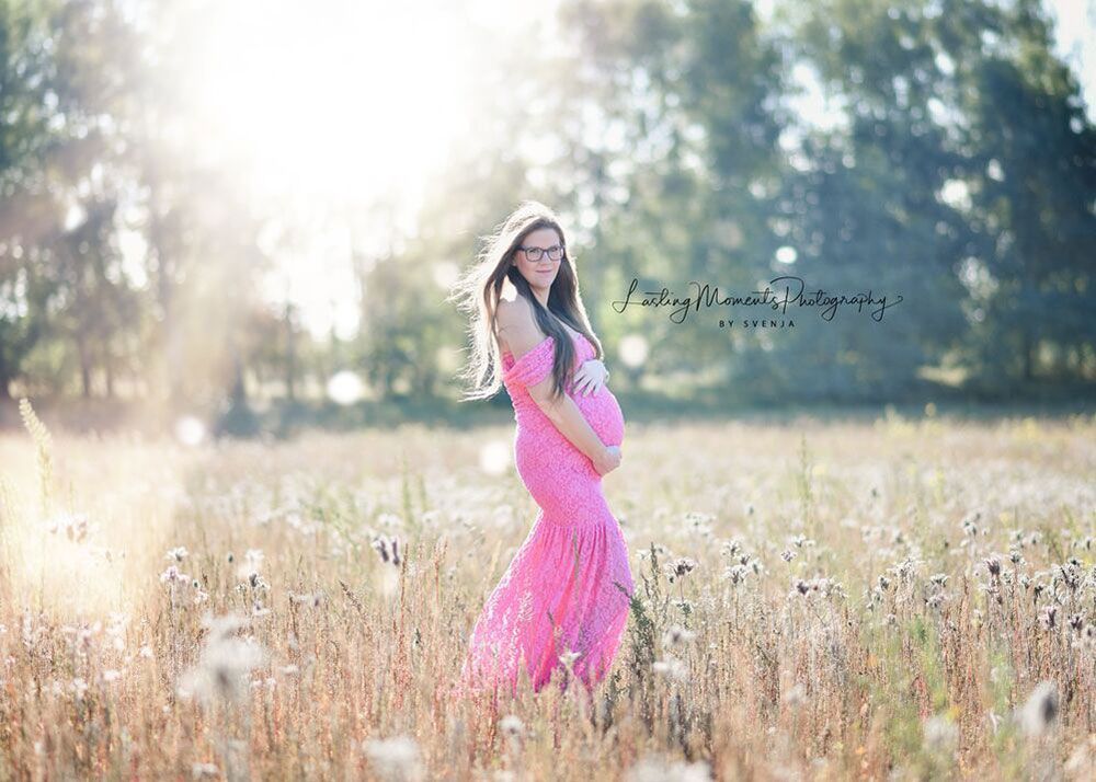 Eine schwangere Frau in einem rosa Kleid steht auf einem Feld und hält ihren Bauch.