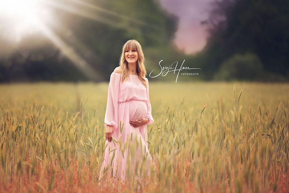 Eine schwangere Frau in einem rosa Kleid steht auf einem Feld und hält ihren Bauch.