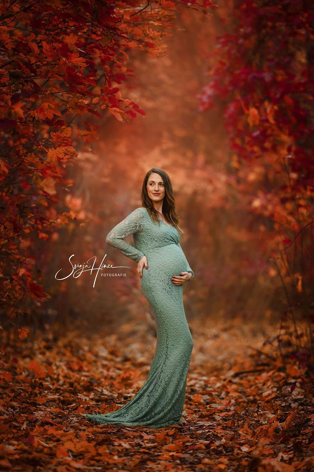 Eine schwangere Frau in einem grünen Kleid steht in einem Wald, umgeben von Blättern.