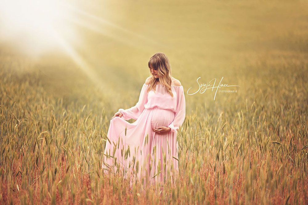 Eine schwangere Frau in einem rosa Kleid steht in einem Weizenfeld.