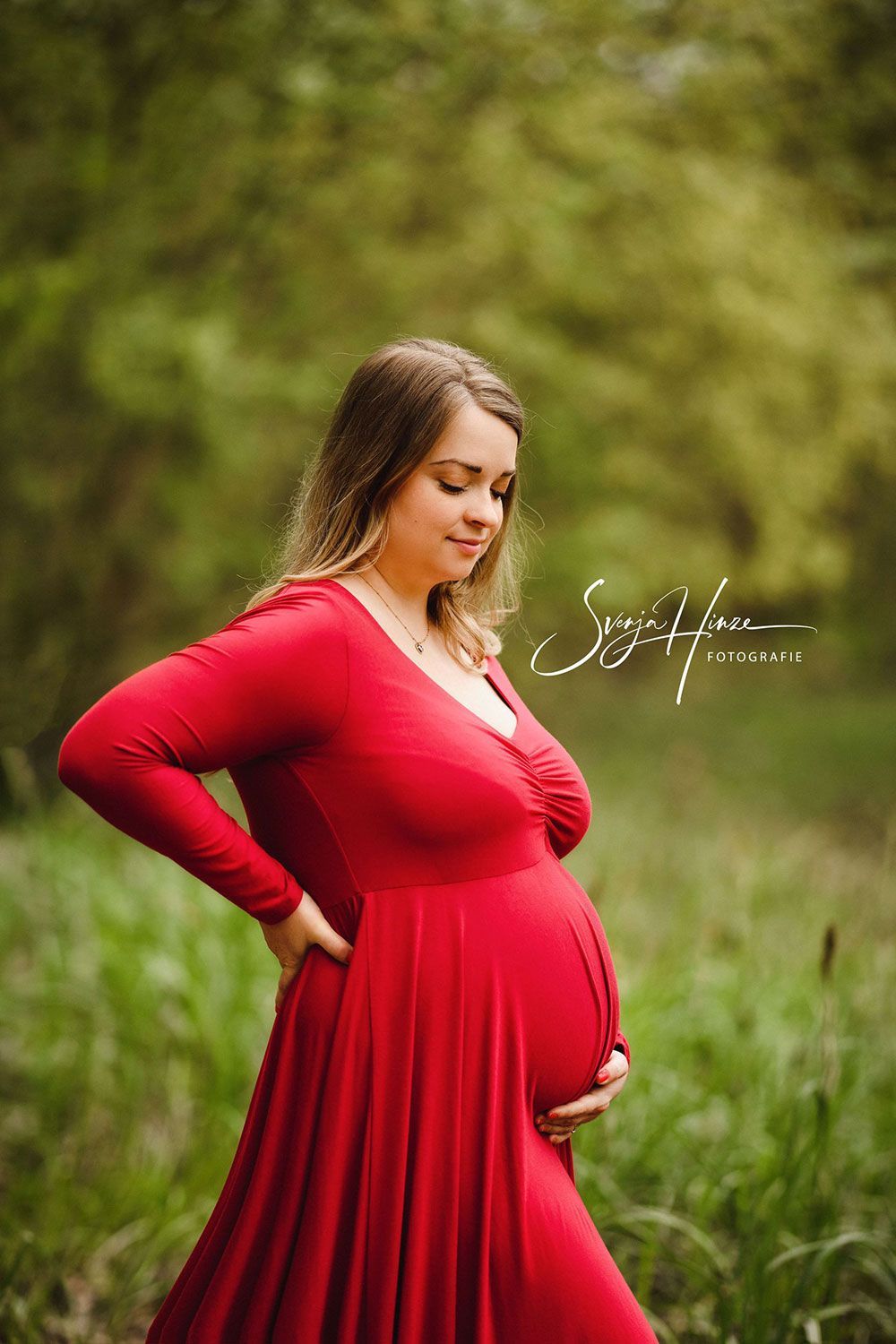 Eine schwangere Frau in einem roten Kleid steht auf einem Feld und hält ihren Bauch.