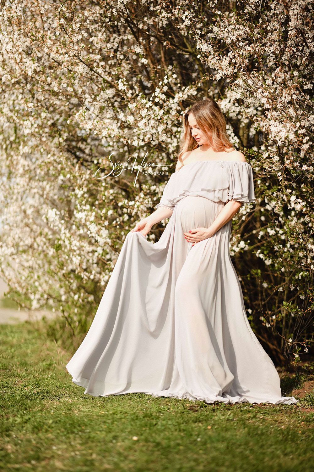 Eine schwangere Frau in einem langen weißen Kleid steht vor einem blühenden Baum.