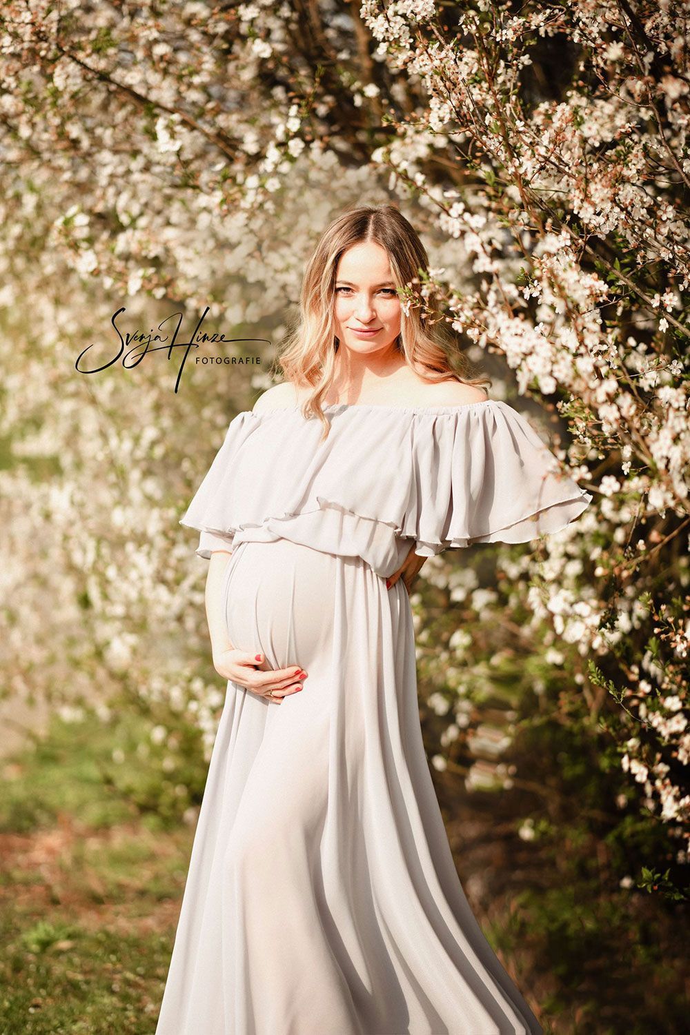 Eine schwangere Frau in einem weißen Kleid steht vor einem blühenden Baum.