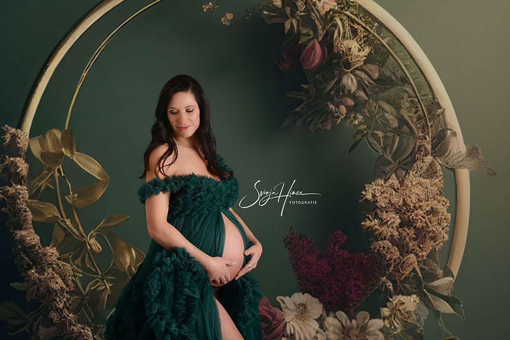 Eine schwangere Frau in einem grünen Kleid steht vor einem Blumenkranz.