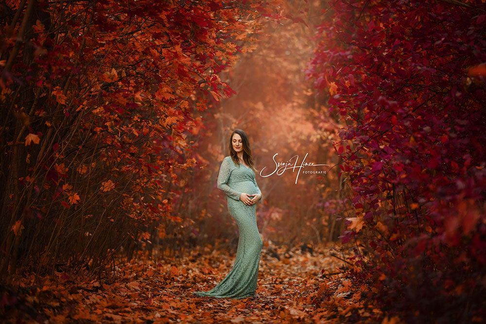 Eine schwangere Frau in einem grünen Kleid steht in einem Wald, umgeben von roten Blättern.