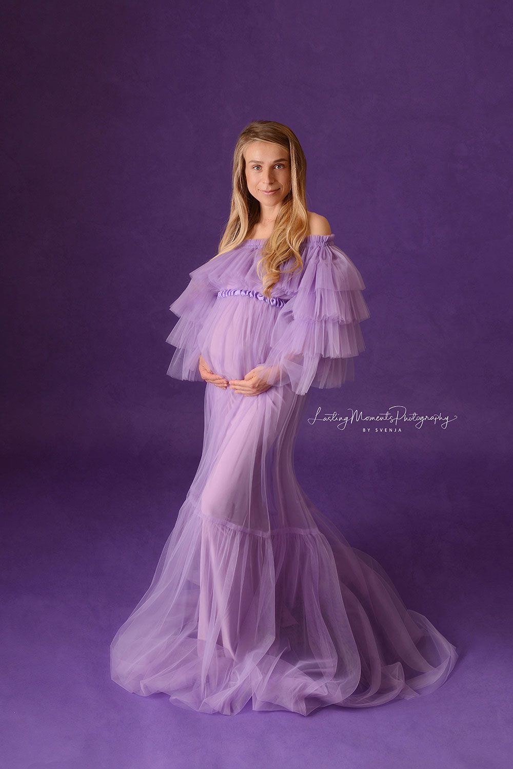 Eine schwangere Frau in einem lila Kleid steht vor einem lila Hintergrund.