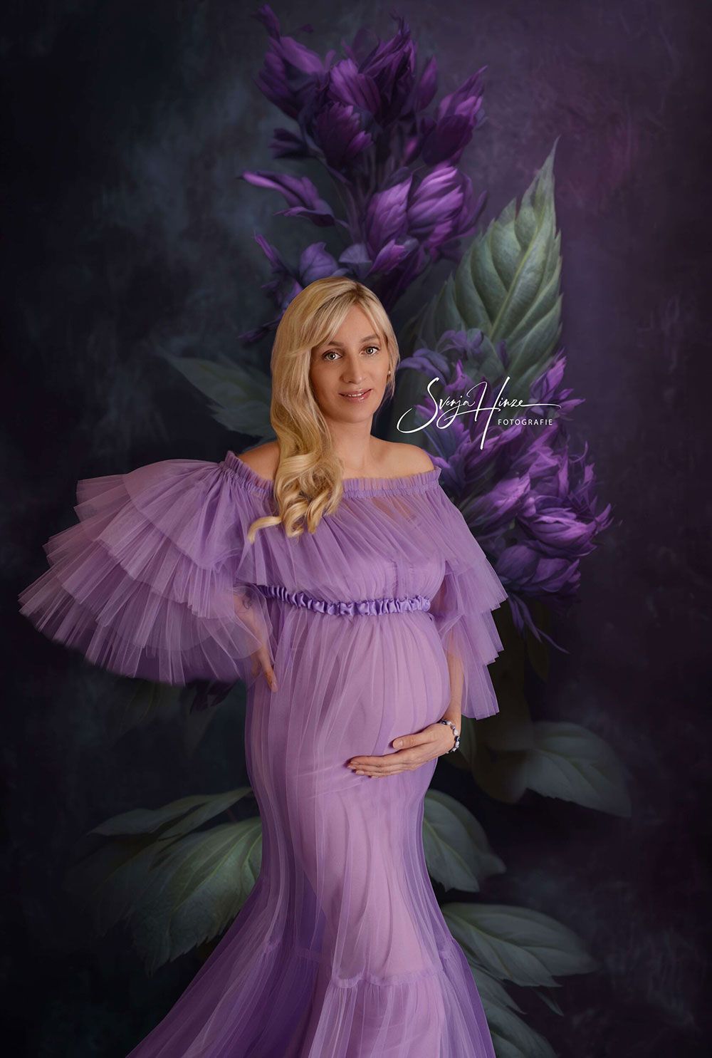 Eine schwangere Frau in einem lila Kleid steht vor lila Blumen.