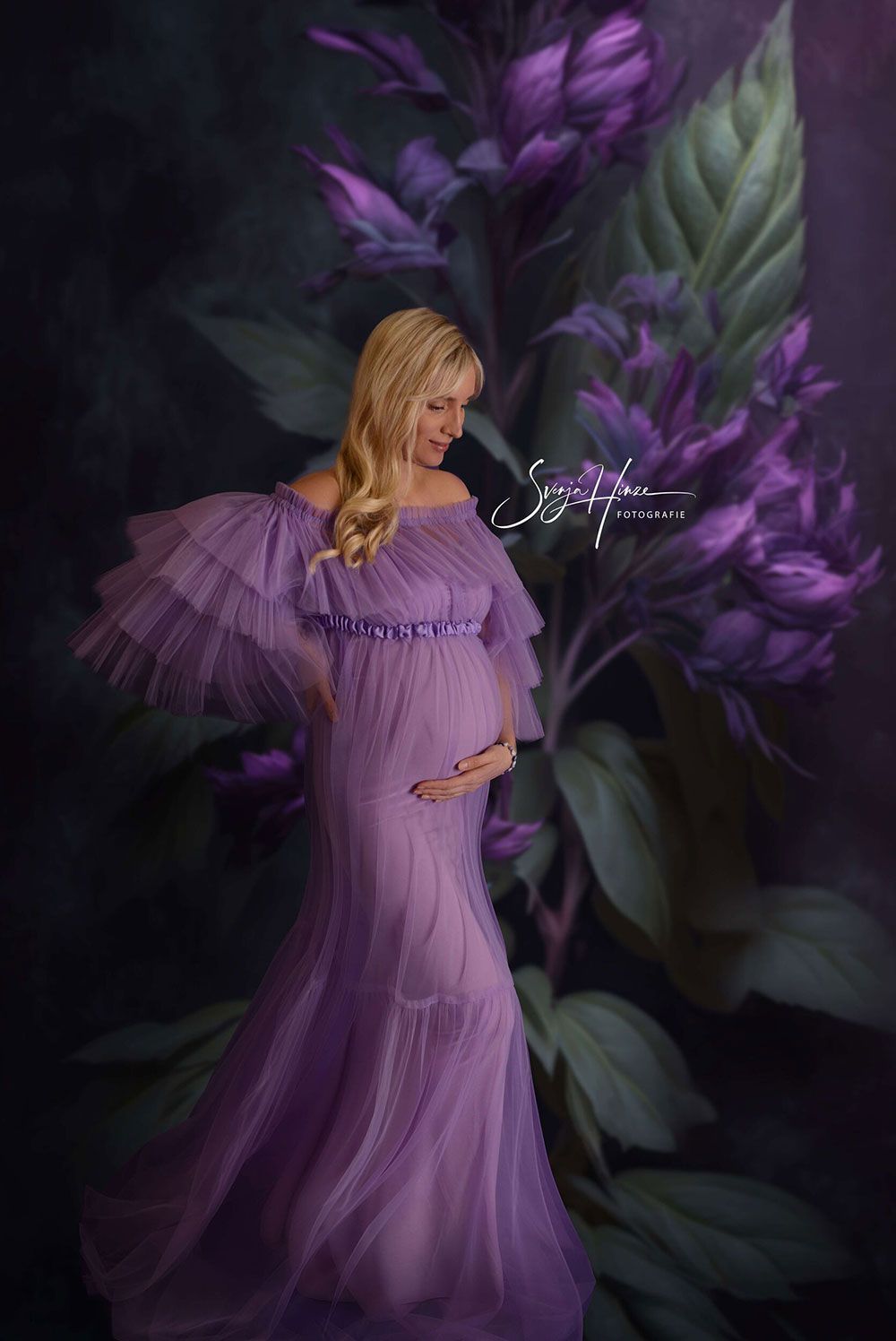 Eine schwangere Frau in einem lila Kleid steht vor lila Blumen.