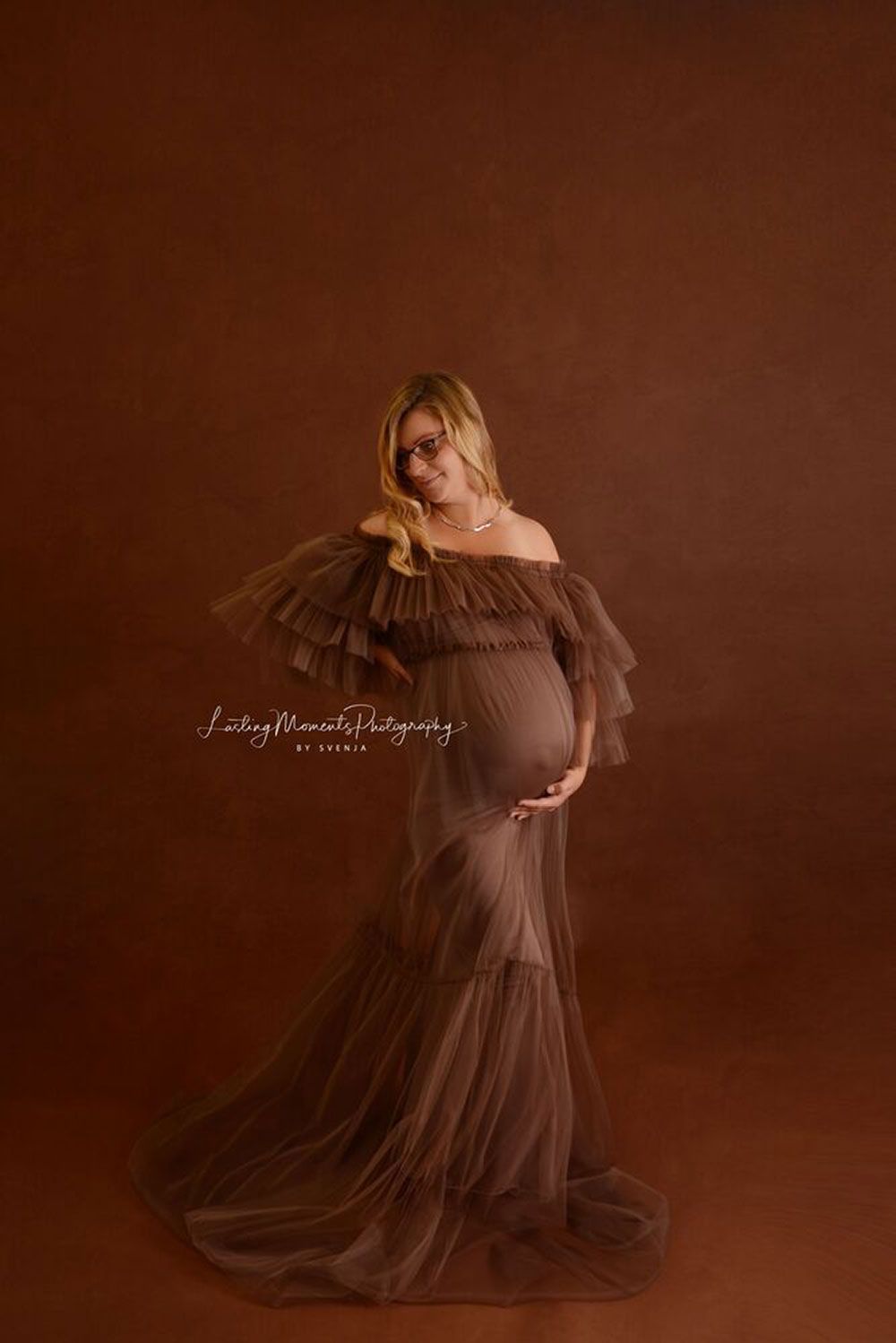 Eine schwangere Frau in einem langen braunen Kleid steht vor einem braunen Hintergrund.