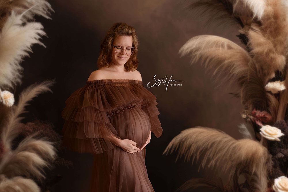 Eine schwangere Frau in einem braunen Kleid steht vor einer Wand aus Federn.