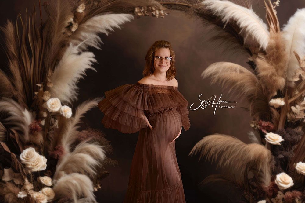Eine schwangere Frau in einem braunen Kleid steht vor einem Federkranz.