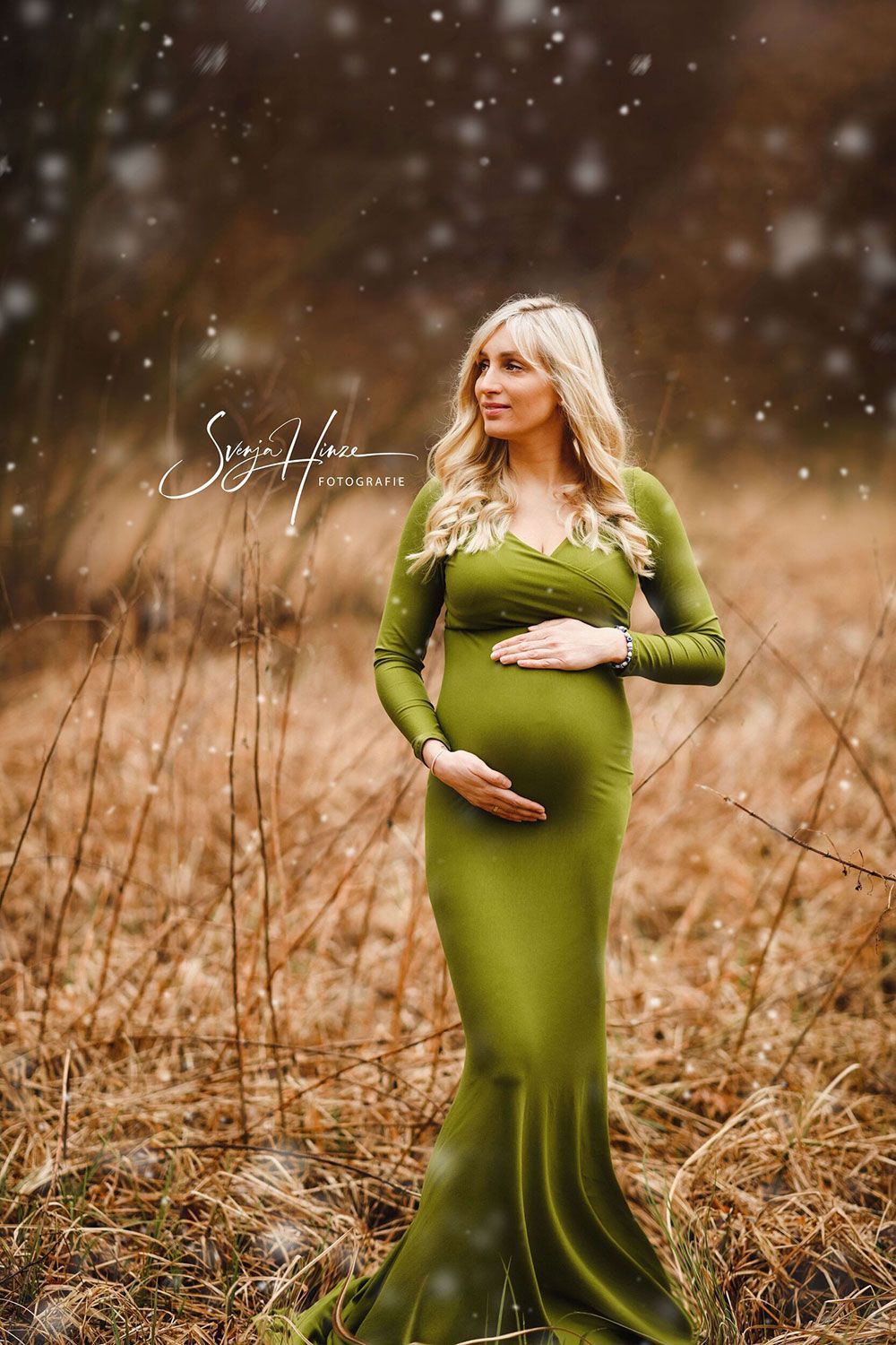 Eine schwangere Frau in einem grünen Kleid steht auf einem Feld und hält ihren Bauch.