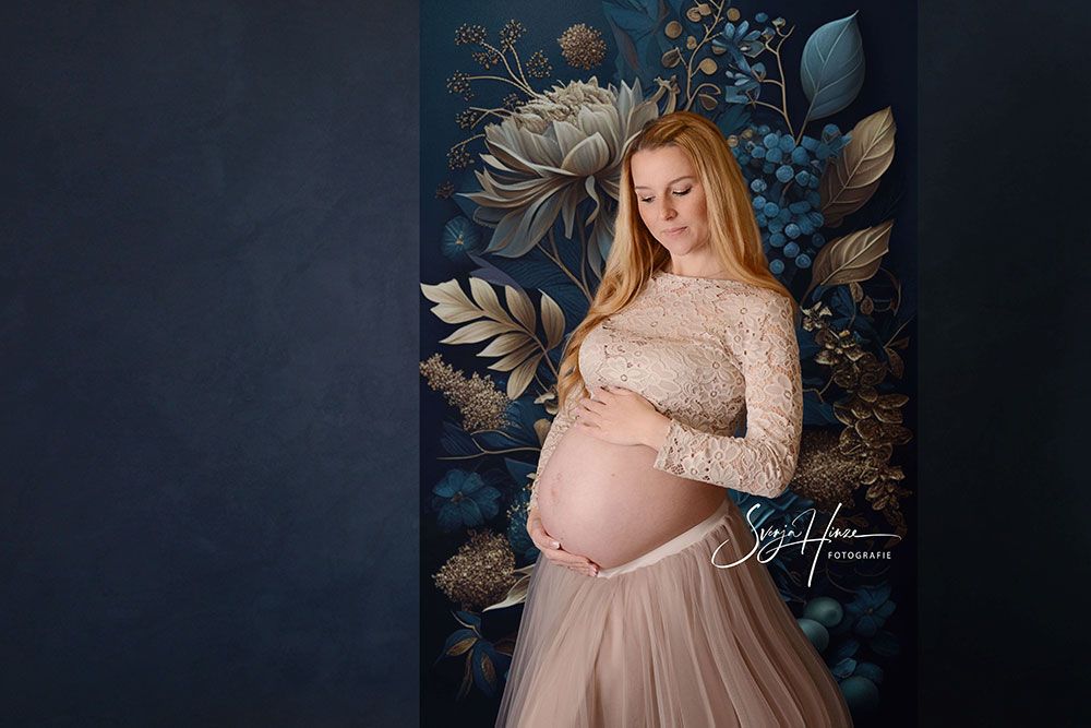 Eine schwangere Frau steht vor einer Blumenwand und hält ihren Bauch.