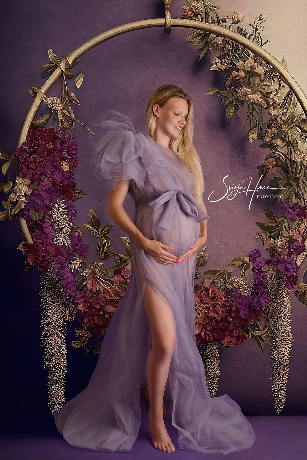 Eine schwangere Frau in einem lila Kleid steht vor einem Blumenbogen.