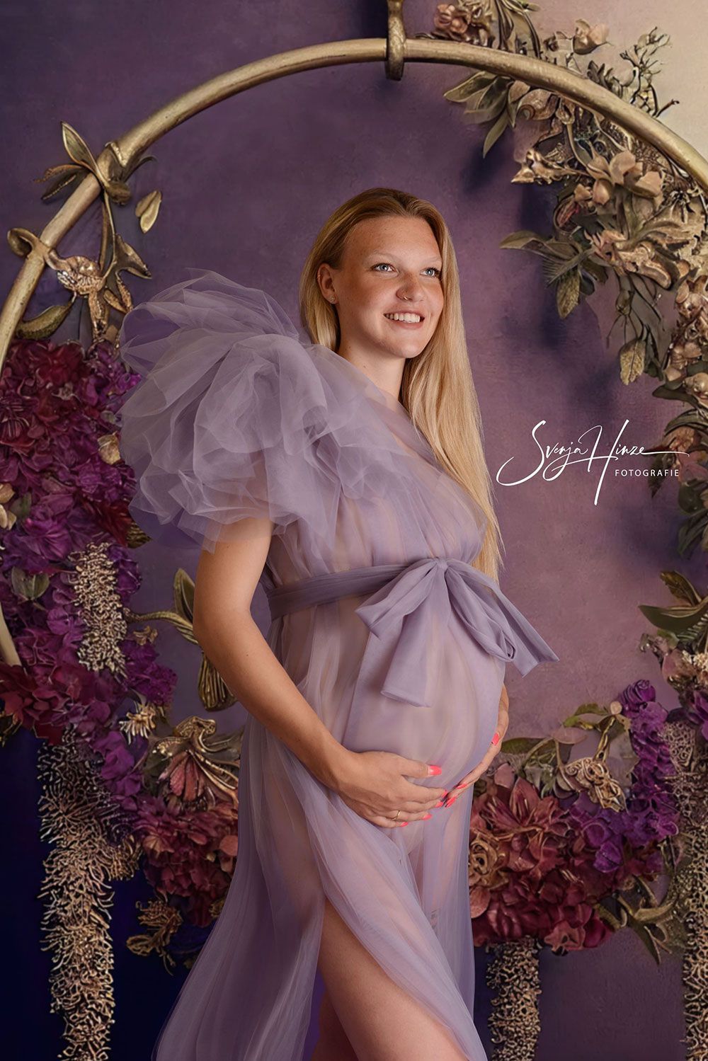 Eine schwangere Frau in einem lila Kleid steht vor lila Blumen.