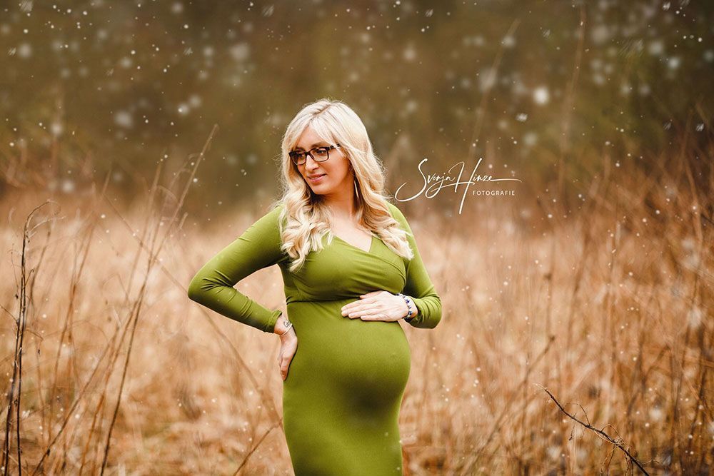 Eine schwangere Frau in einem grünen Kleid steht auf einem Feld und hält ihren Bauch.