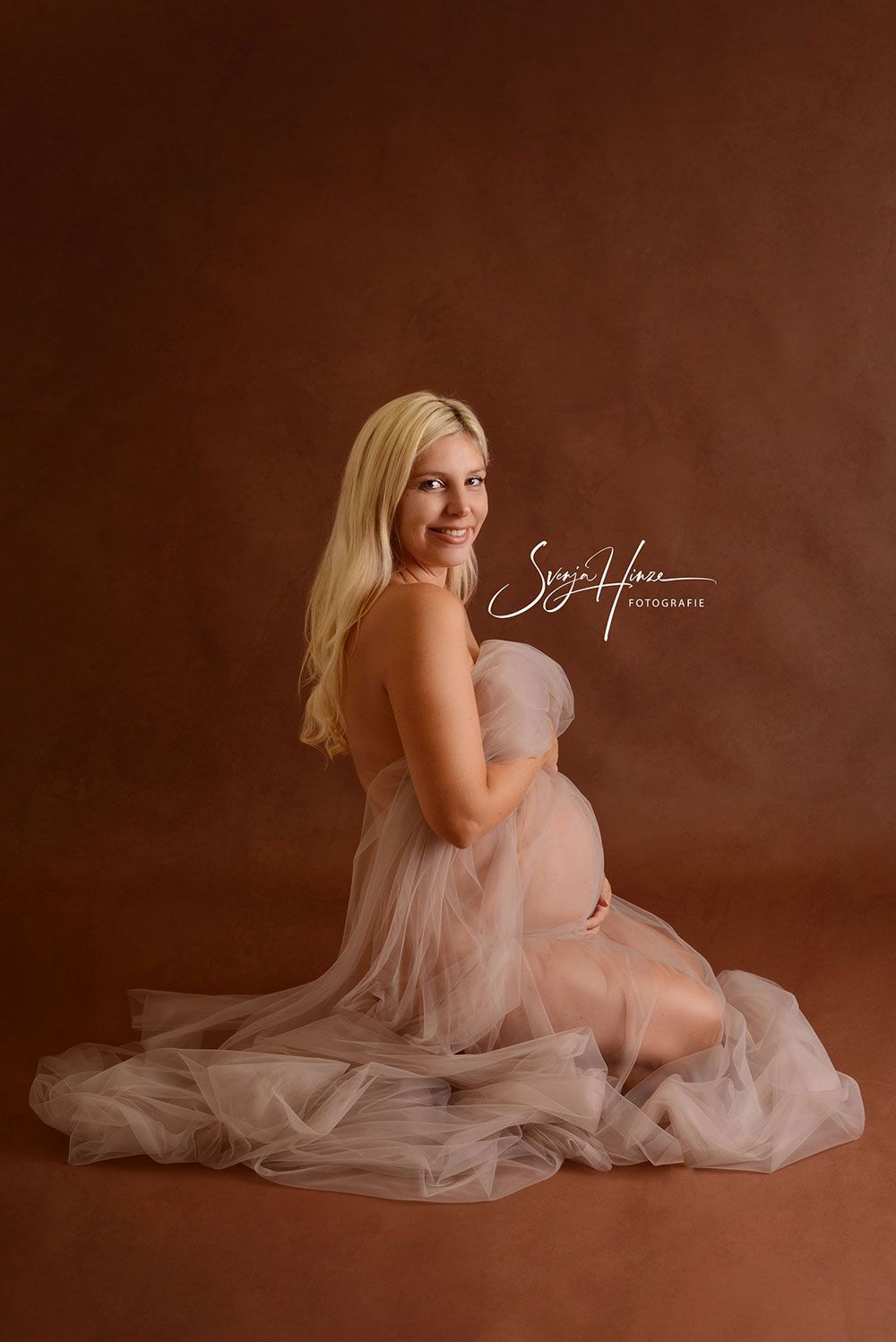 Eine schwangere Frau sitzt in einem Tüllkleid auf dem Boden.