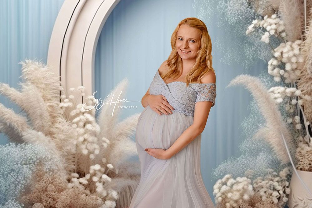 Eine schwangere Frau in einem blauen Kleid steht vor einem Fenster und hält ihren Bauch.