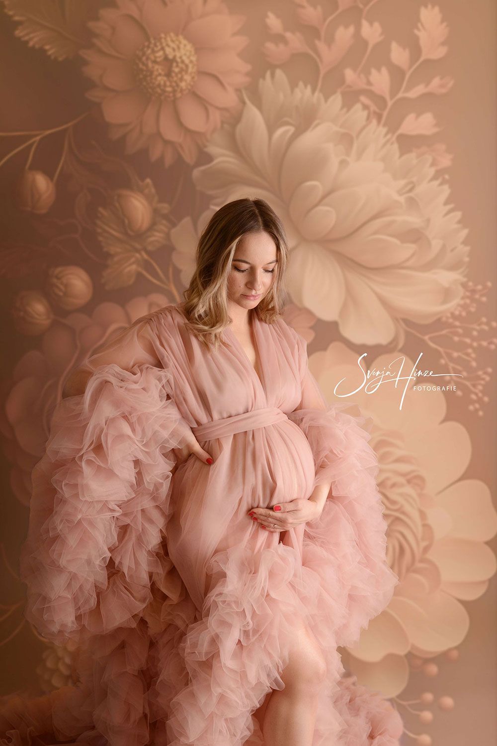 Eine schwangere Frau in einem rosa Kleid steht vor einer Blumenwand.