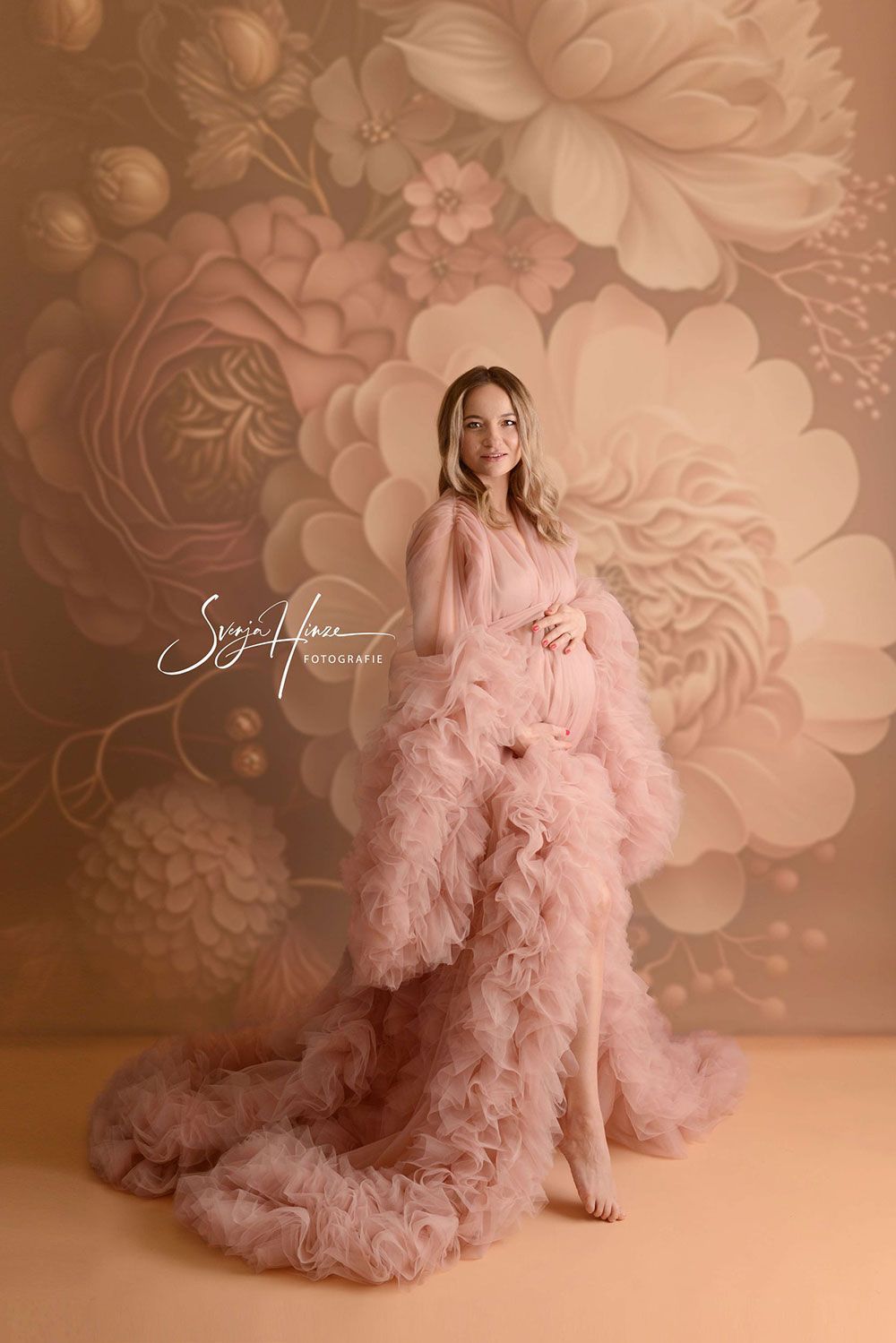 Eine schwangere Frau in einem langen rosa Kleid steht vor einer großen Blumenwand.