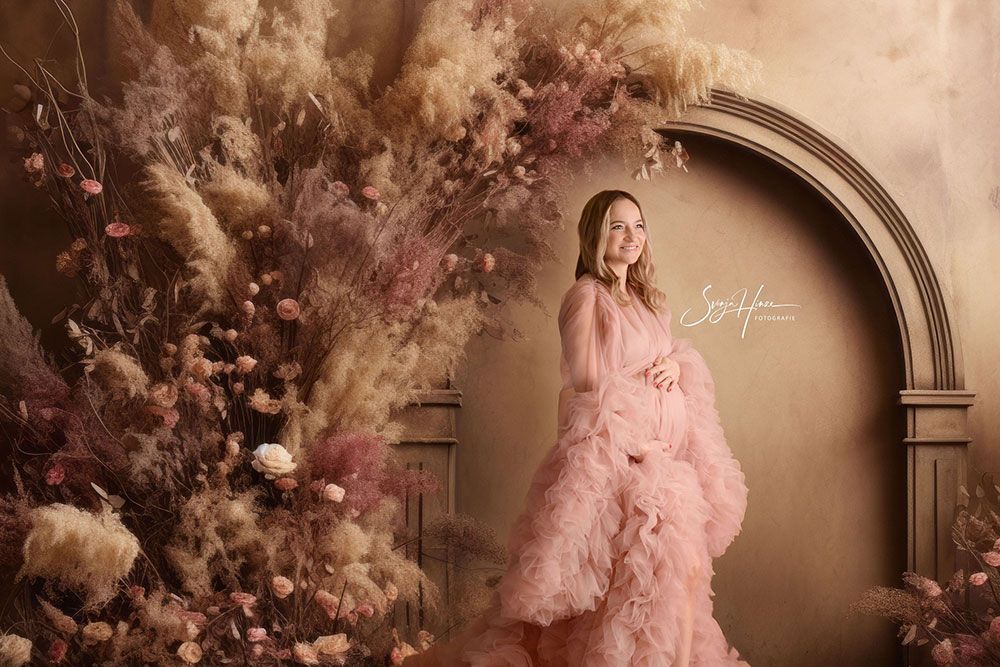 Eine schwangere Frau in einem rosa Kleid steht vor einer Wand aus getrockneten Blumen.