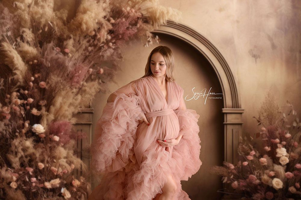 Eine schwangere Frau in einem rosa Kleid steht vor einer Wand mit Blumen.