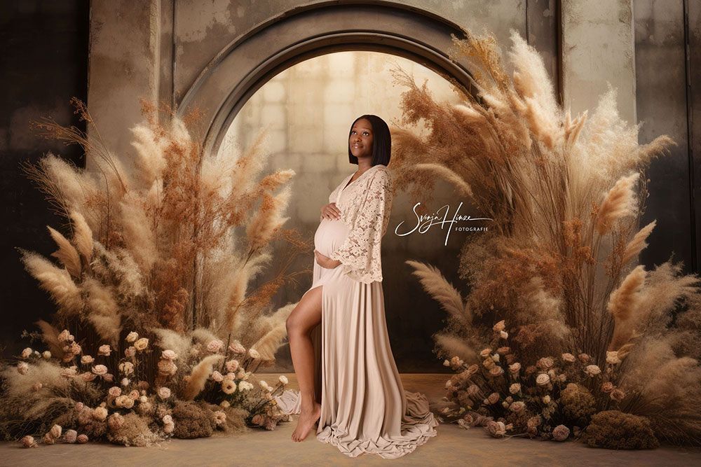 Eine schwangere Frau steht vor einer Mauer, die von hohem Gras umgeben ist.