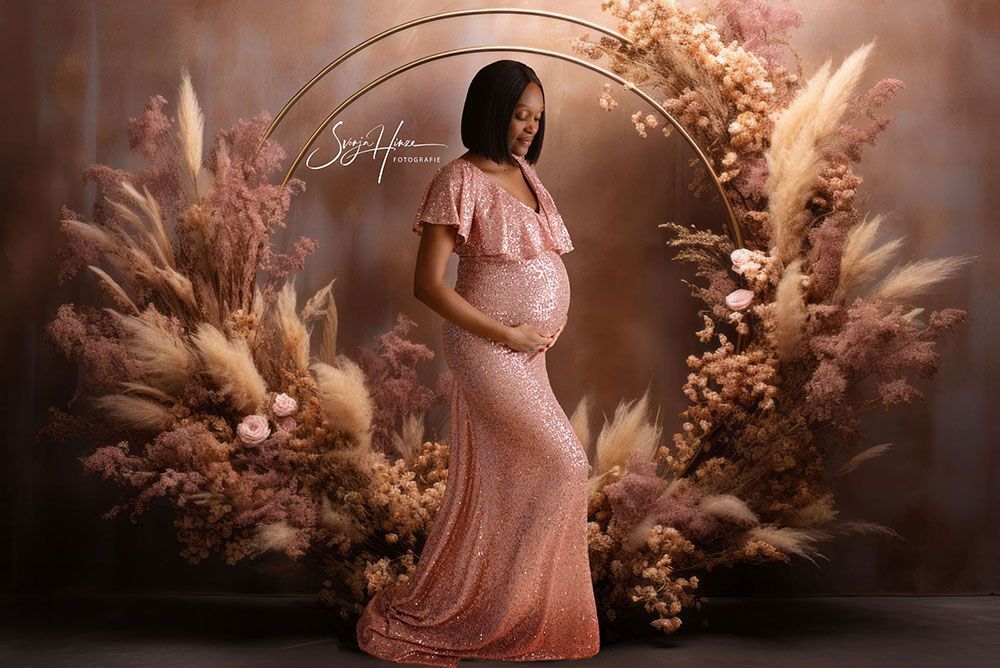 Eine schwangere Frau in einem rosa Kleid steht vor einem Blumenbogen.