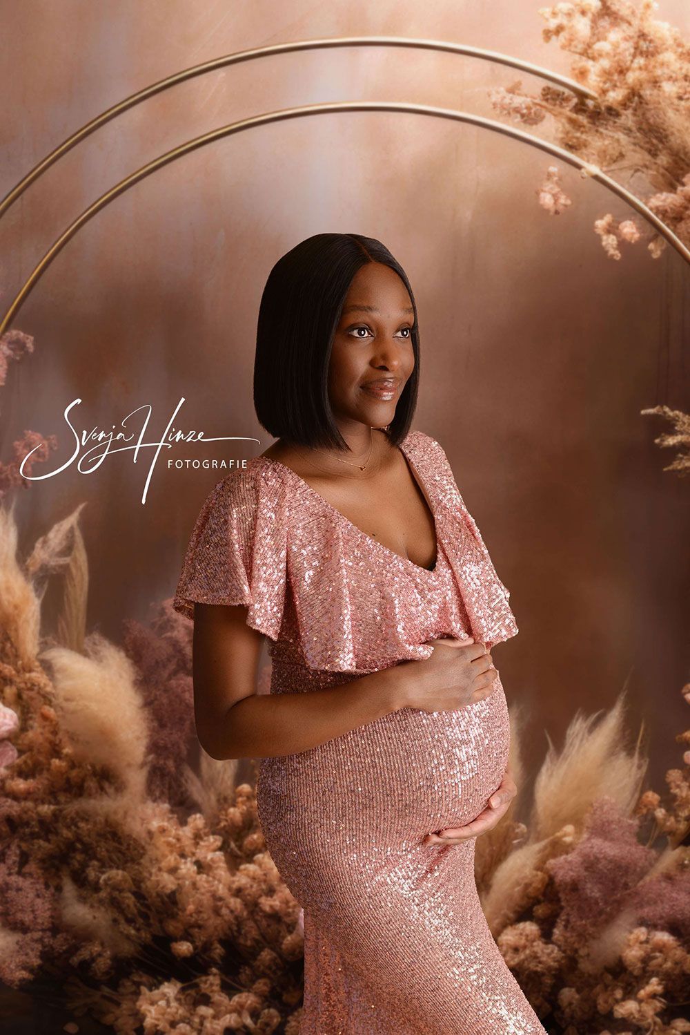 Eine schwangere Frau in einem rosa Paillettenkleid hält ihren Bauch.