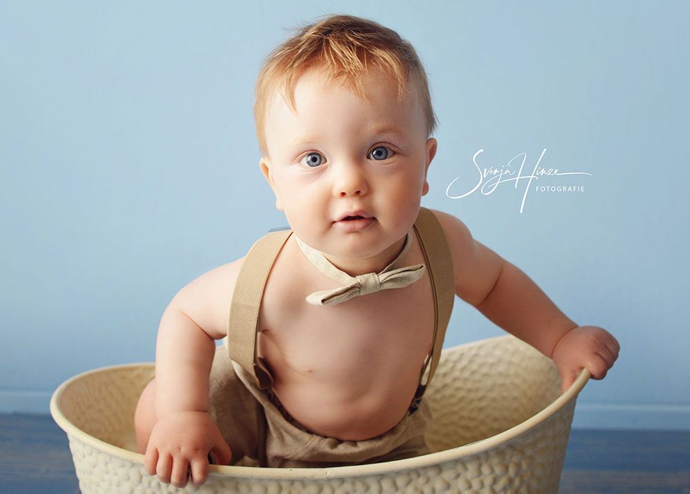 Ein Baby sitzt mit Hosenträgern und Fliege in einem Korb.