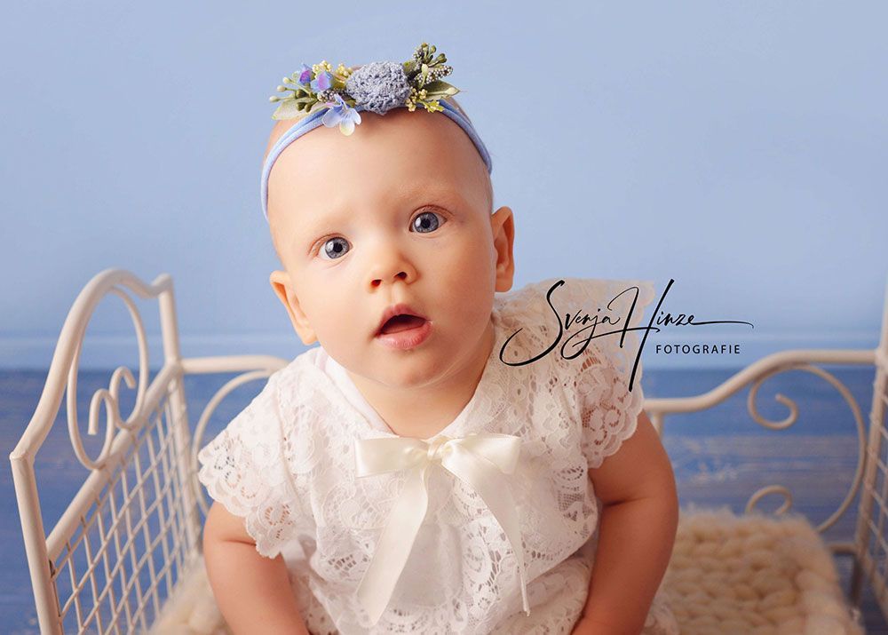 Ein kleines Mädchen mit einem weißen Kleid und einem Stirnband sitzt in einem weißen Kinderbett.