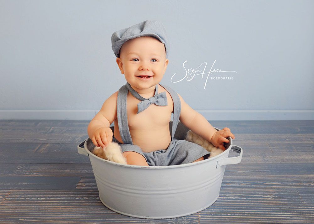 Ein Baby sitzt mit Hosenträgern und Mütze in einem Eimer.