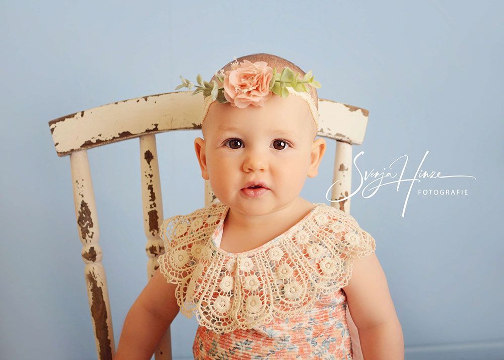 Ein kleines Mädchen sitzt mit einem Blumenkranz auf dem Kopf auf einem Stuhl.