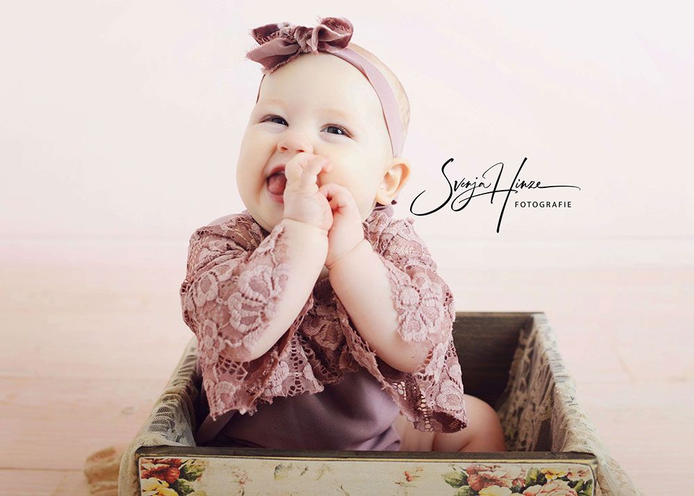 Ein Baby sitzt in einer Kiste und hat die Hand auf der Nase.