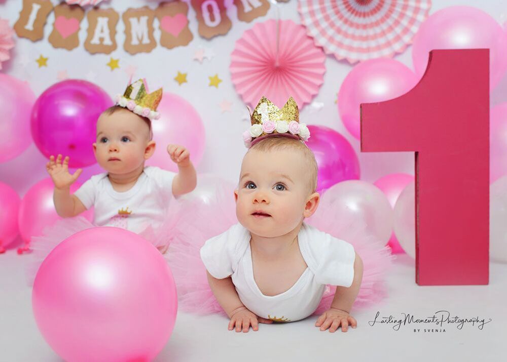Zwei Babys mit Kronen sitzen mit rosa Luftballons auf dem Boden.