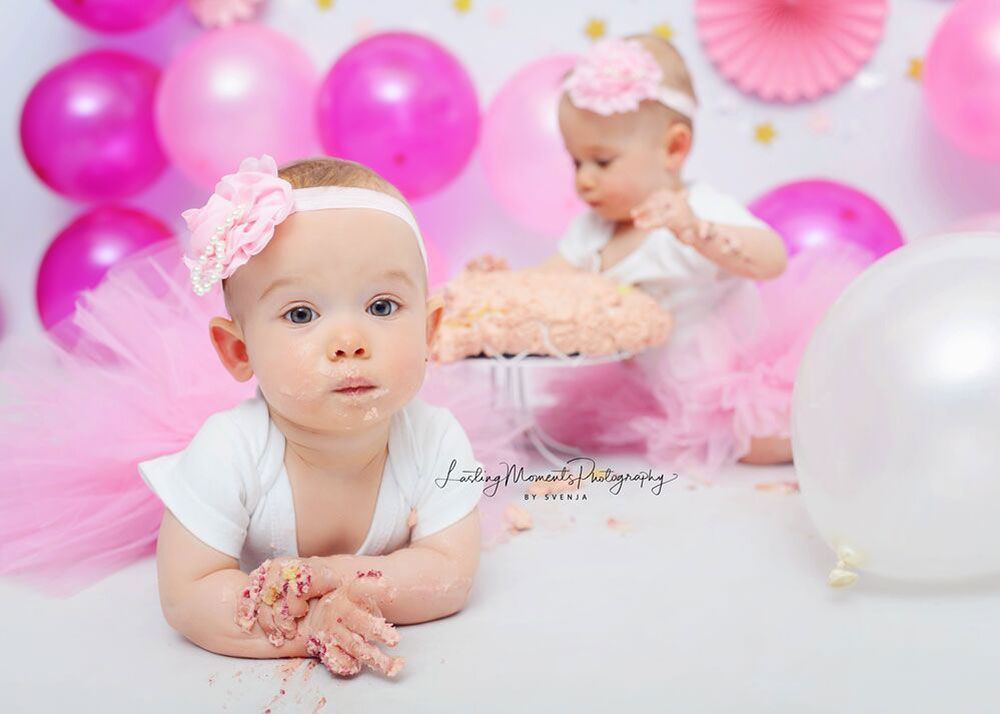 Zwei kleine Mädchen sitzen nebeneinander vor einem Kuchen und Luftballons.