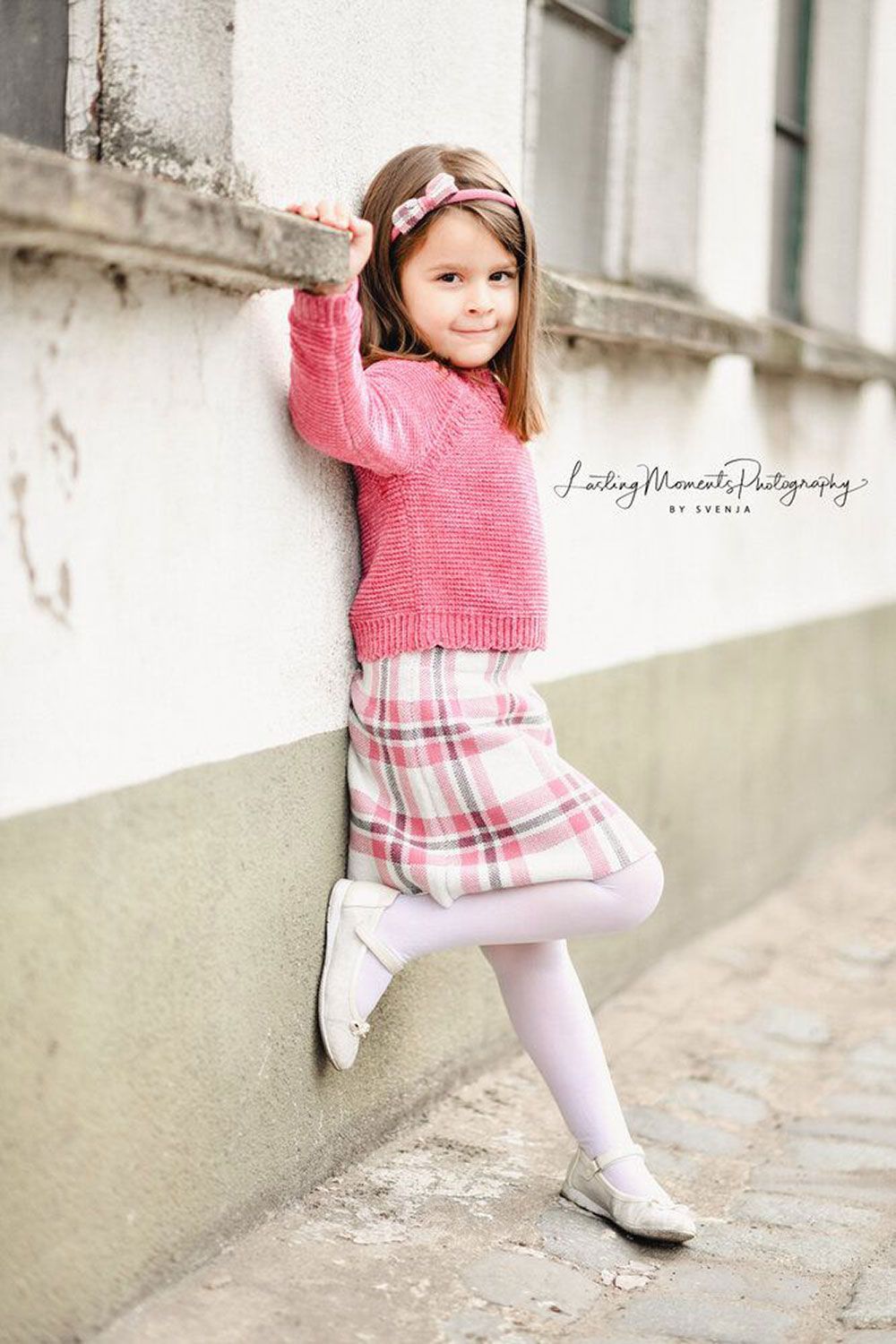 Ein kleines Mädchen in einem rosa Pullover und einem karierten Rock lehnt an einer Wand.