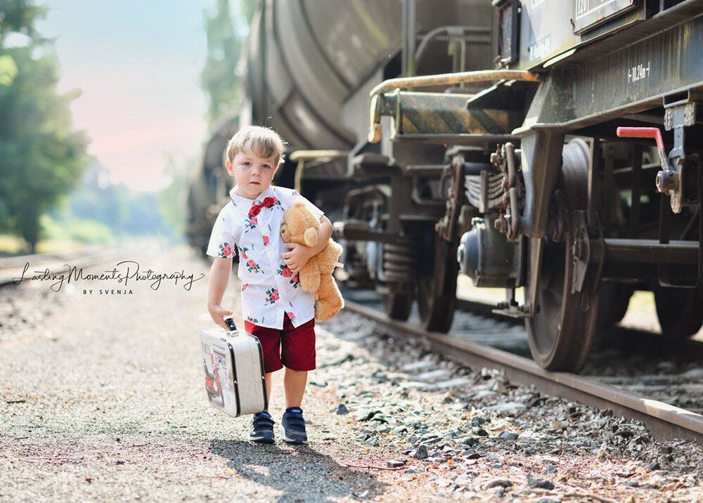 Ein kleiner Junge hält einen Teddybären und einen Koffer auf Bahngleisen.