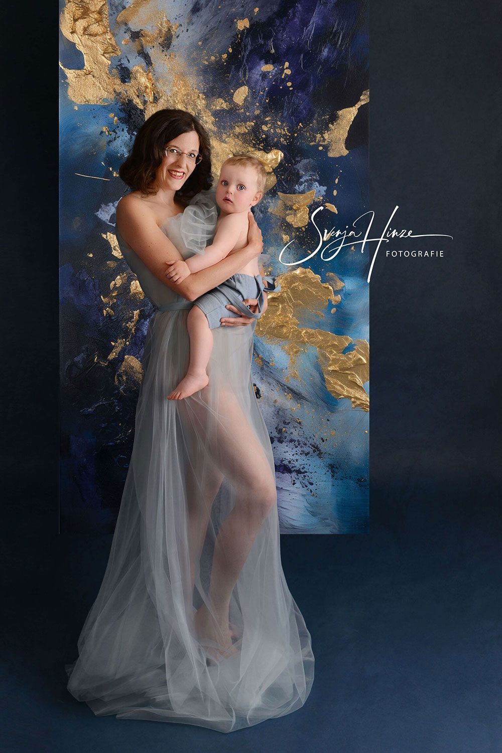 Eine Frau in einem langen Kleid hält ein Baby vor einem Gemälde.