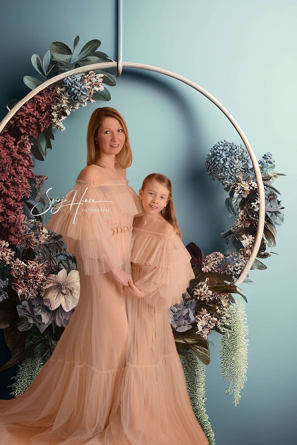 Eine schwangere Frau und ein kleines Mädchen stehen nebeneinander vor einem Blumenkranz.