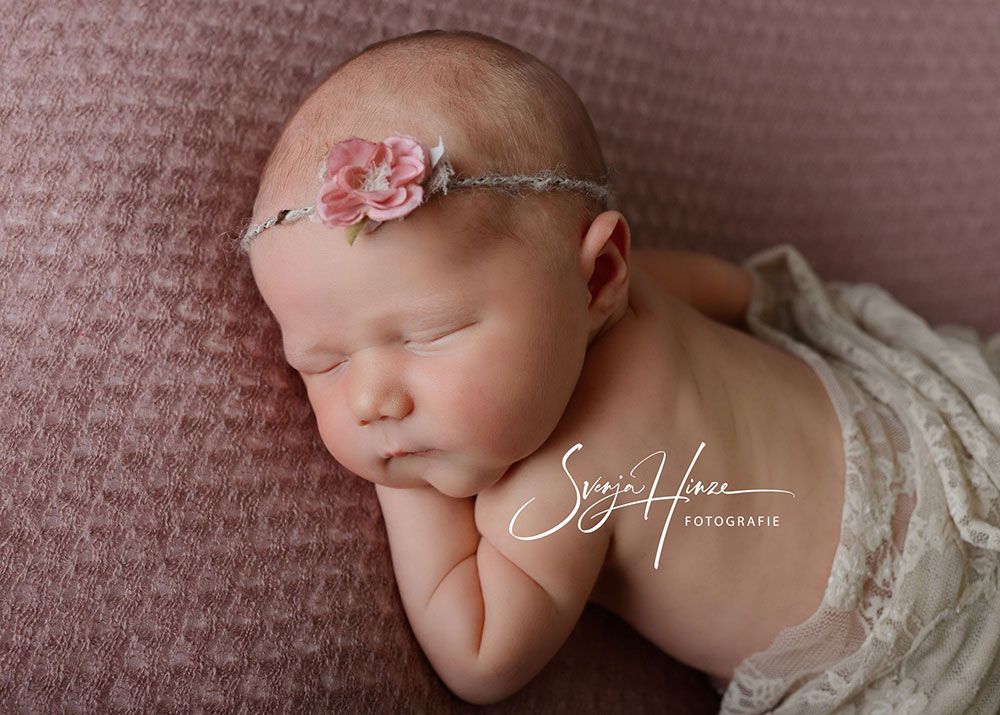 Ein Neugeborenes schläft mit einem Blumenstirnband auf einer rosa Decke.