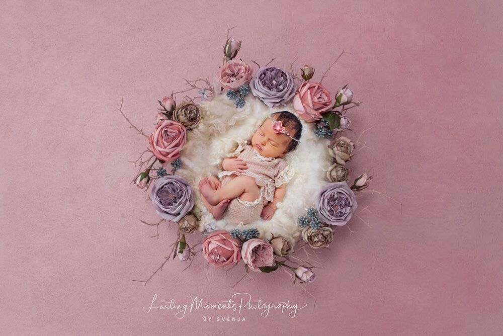 Ein Neugeborenes liegt in einem Blumenkranz auf rosa Hintergrund.