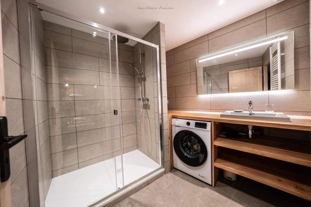Salle de bains moderne avec douche en verre, laveuse, comptoir en bois et miroir éclairé.